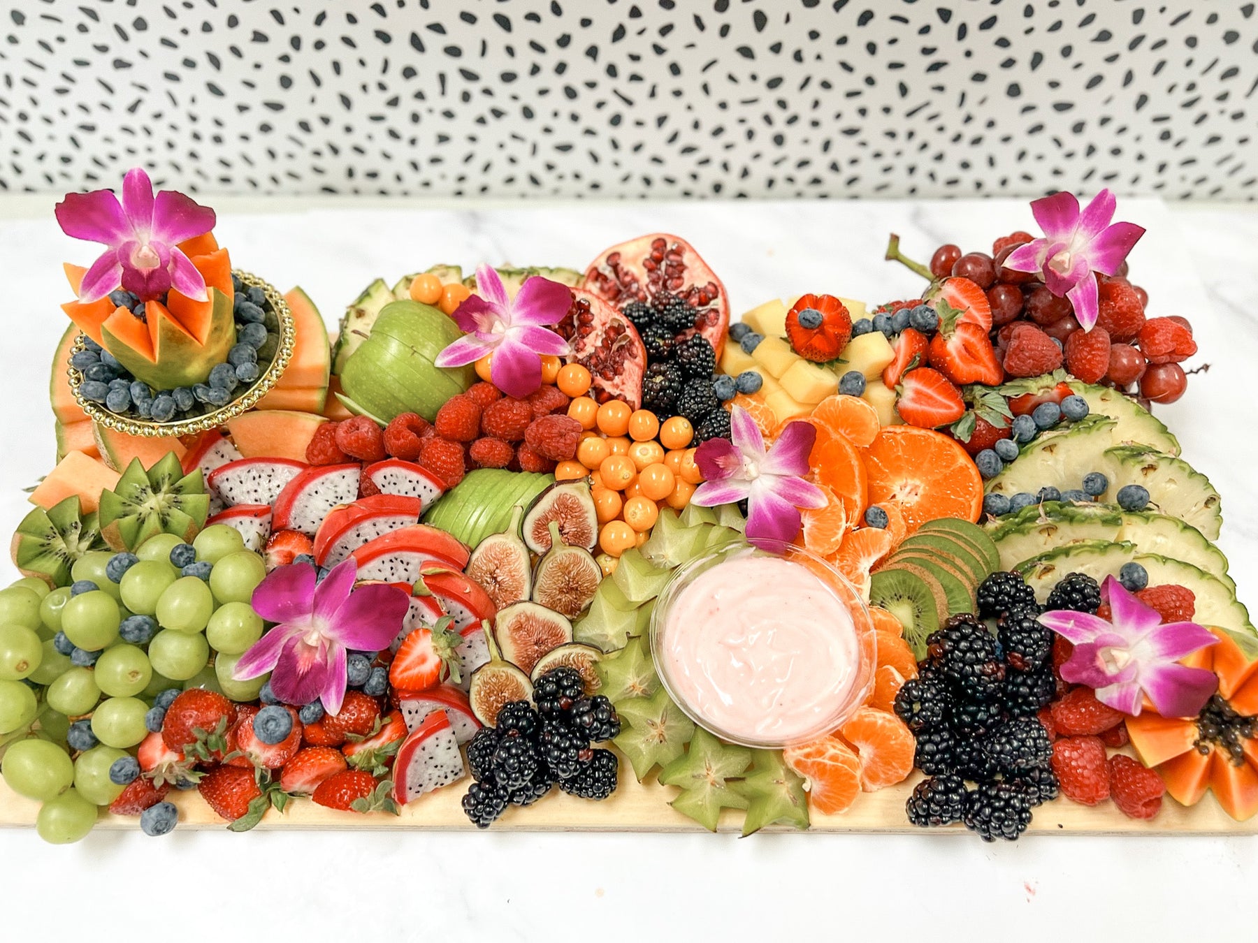 An assortment of fresh fruits arranged on a board, including berries, citrus, and possibly a dragon fruit, garnished with flowers and accompanied by a pink dip.