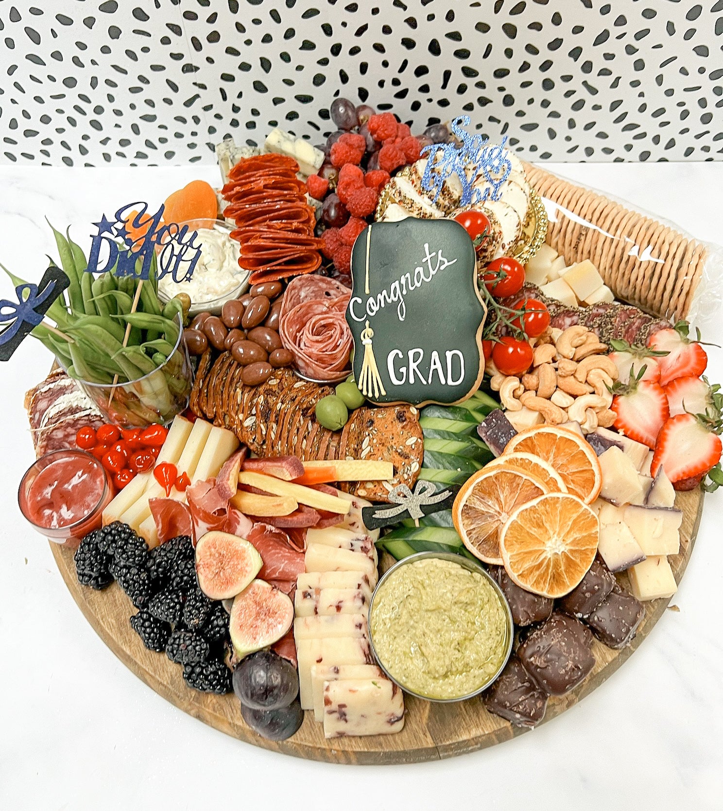 A charcuterie board with an assortment of meats, cheeses, fruits, and vegetables, topped with a sign that reads 'Congrats Grad'.